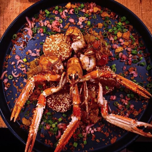 A vibrant seafood dish featuring a large crab, shrimp, and garnished with green onions and crispy toppings on a black plate.