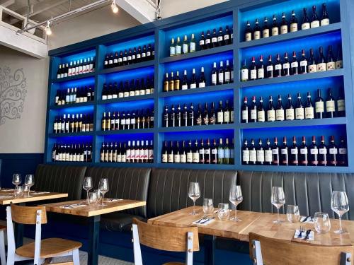 A restaurant interior featuring a blue-lit wall of wine bottles and wooden tables set with glasses and utensils.