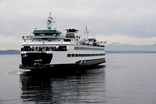 A large ferry sails across calm waters with mountains in the background under a cloudy sky.