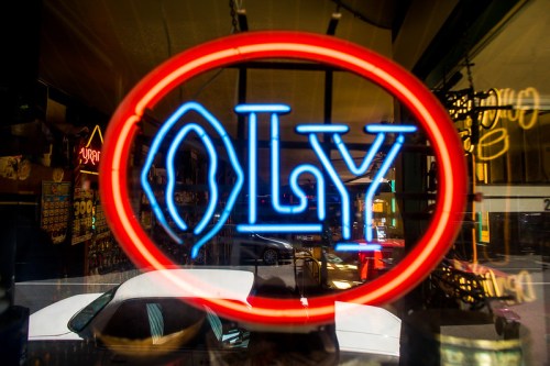 A bright neon sign reading "OLY" in red and blue, reflecting in a window with a car visible inside.