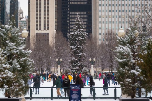 A bustling ice skating rink surrounded by snow-covered trees and tall buildings, with people enjoying winter activities.