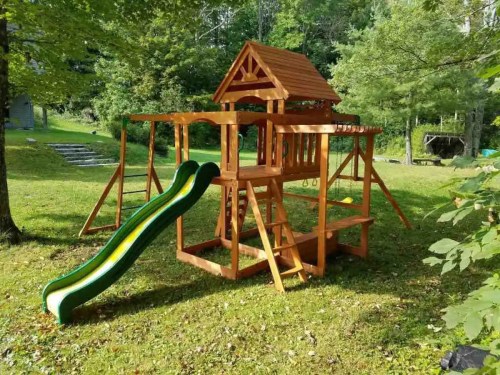 A wooden playground set with a slide, swings, and a playhouse, surrounded by green grass and trees.