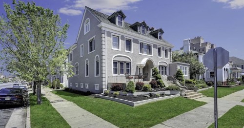 A large, elegant gray house with a manicured lawn and trees, set against a clear blue sky.
