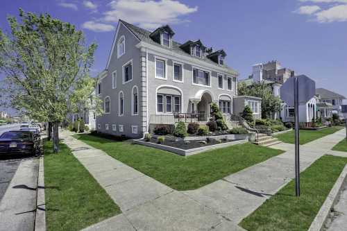 A large, elegant gray house with a manicured lawn and trees, set on a quiet street under a clear blue sky.