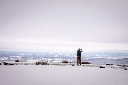 A person stands on a snowy landscape, gazing at a distant view of rolling hills and a cloudy sky.