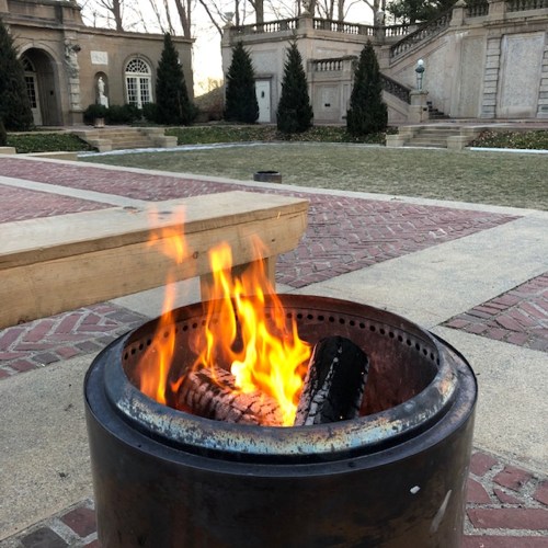 A fire burns in a metal fire pit, surrounded by a brick patio and landscaped gardens in the background.