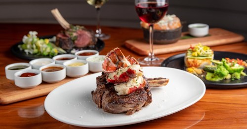 A gourmet steak topped with lobster, surrounded by sauces, with wine and other dishes in the background.