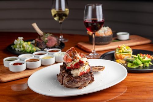 A gourmet steak topped with lobster, served on a white plate, with wine glasses and various dishes in the background.
