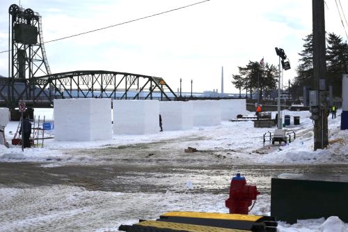 Snow blocks line a snowy area near a river, with a bridge in the background and a cloudy sky above.