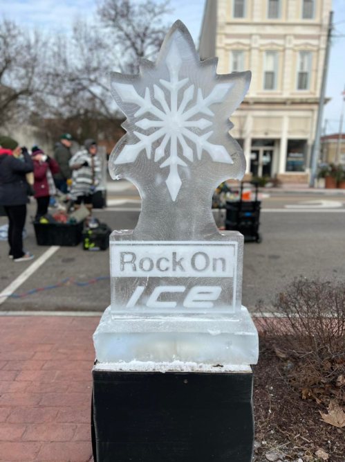 Ice sculpture of a snowflake with the text "Rock On Ice" at a winter event, with people in the background.