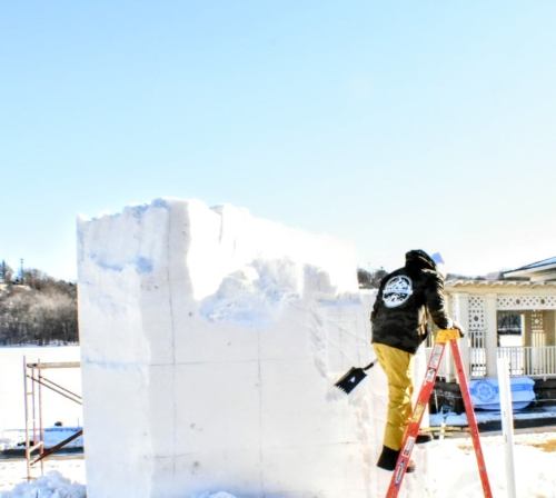 A person in yellow pants uses a shovel to shape a large snow block near a frozen lake and a house in the background.