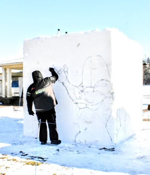 A person in a black jacket sketches on a large snow block, with a snowy landscape in the background.
