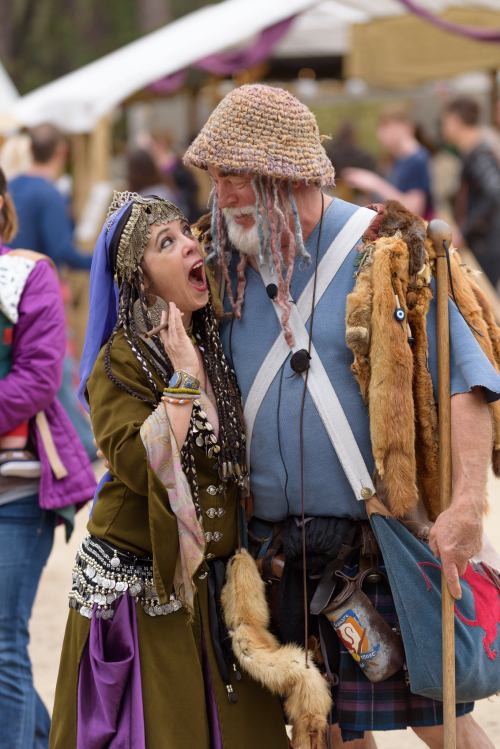 A woman in a colorful costume playfully interacts with a man in a kilt and hat at a lively outdoor festival.