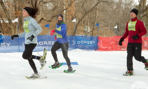 Three participants in snowshoes run through a snowy course, surrounded by trees and colorful banners.