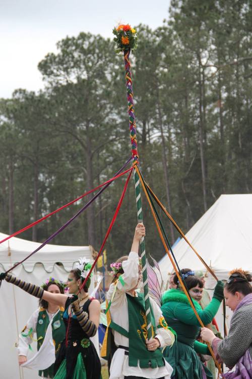 A group of people celebrate with a maypole, adorned with colorful ribbons, in a forested outdoor setting.