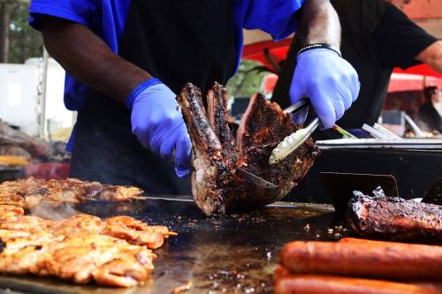 A person in blue gloves slices grilled meat on a barbecue grill, surrounded by various cooked meats.