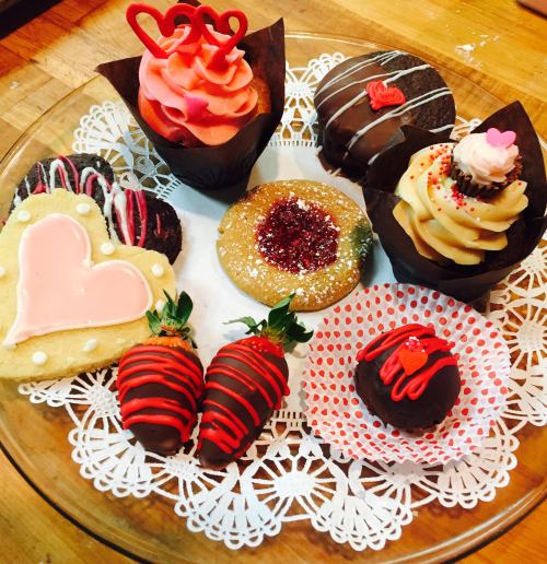 A platter of decorated sweets, including cupcakes, cookies, and chocolate-covered strawberries, arranged on lace.