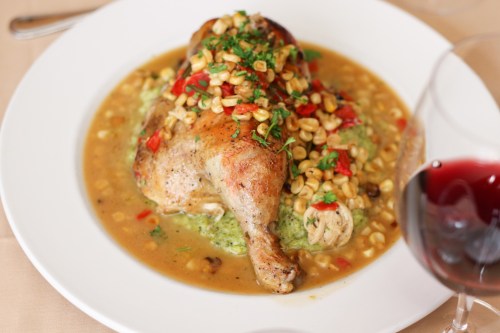 A plated dish featuring roasted chicken topped with corn salsa, served on a bed of green puree, with a glass of red wine.