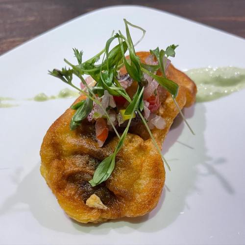 A golden fried dish topped with fresh herbs and diced vegetables, served on a white plate with a green sauce.