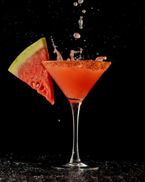 A vibrant cocktail with a watermelon slice, splashes of liquid, and a rim of seasoning, set against a black background.