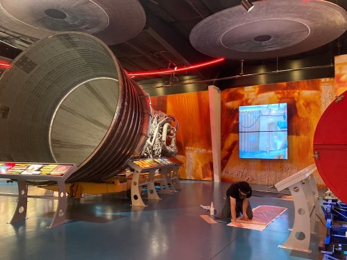 A large rocket engine display in a museum, with a person working on the floor and colorful informational panels nearby.