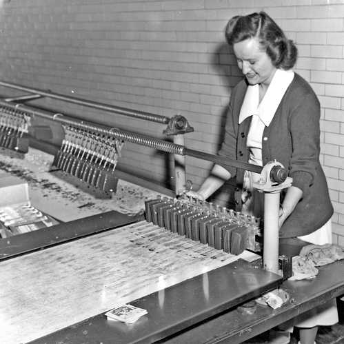 A woman operates a vintage printing press, carefully managing the printing process in a factory setting.