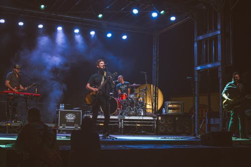A band performs on stage with colorful lights, featuring a singer with a guitar, keyboardist, drummer, and bassist.