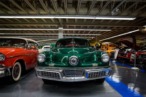 A vintage green car displayed in a museum, surrounded by other classic cars in a well-lit garage.