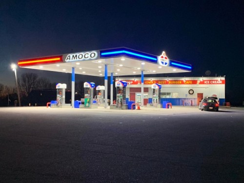A brightly lit Amoco gas station at night, featuring fuel pumps and a diner with a sign for ice cream.