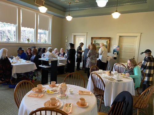 A cozy tea gathering with women seated at tables, enjoying tea and conversation in a warmly lit room.