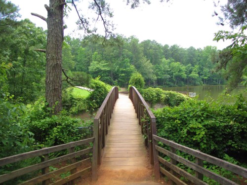 A wooden bridge leads over a calm lake, surrounded by lush green trees and foliage.