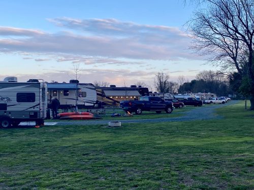 A campsite with several RVs and trucks parked on a grassy area, under a cloudy sky at dusk.