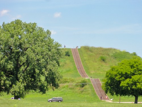 A grassy hill with a sloped path, surrounded by trees and a clear blue sky, with people walking up the incline.