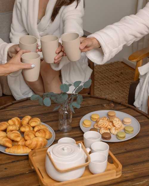 A cozy gathering with friends, toasting with cups, surrounded by pastries and macarons on a wooden table.