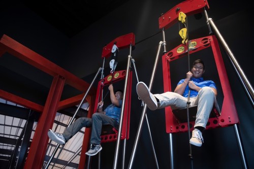 Two people enjoying swings in a modern indoor play area, surrounded by red structures and a dark background.