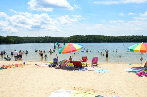 A sunny beach scene with colorful umbrellas, people swimming, and towels spread on the sand by a calm lake.