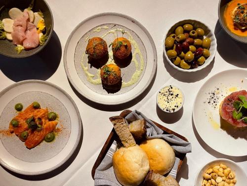 A spread of gourmet dishes including olives, salmon, bread rolls, and small appetizers on a white tablecloth.