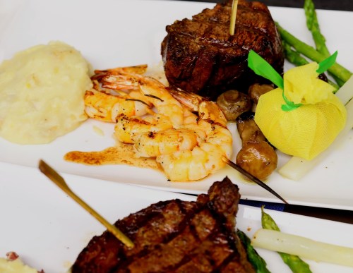 A gourmet plate featuring grilled shrimp, steak, mashed potatoes, and asparagus, garnished with a lemon twist.