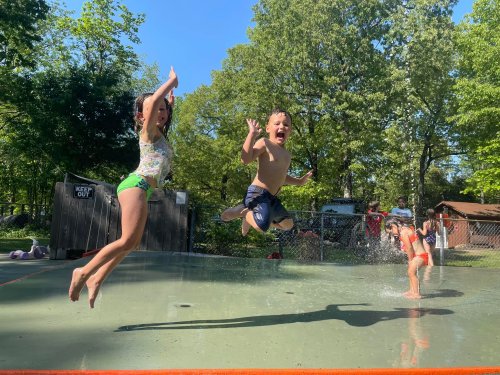 Two children joyfully jumping into a splash pad on a sunny day, with others playing in the background.