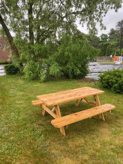 A wooden picnic table and bench sit on a grassy area, surrounded by trees and a rainy atmosphere.