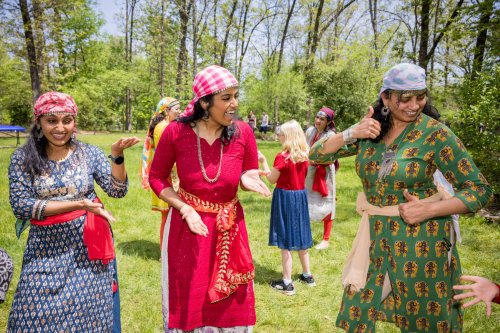 Three women in colorful traditional outfits dance joyfully outdoors, surrounded by greenery and other people in the background.