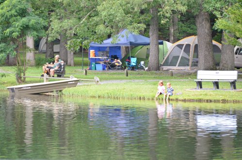 A peaceful lakeside scene with campers, a boat, and children playing by the water's edge among trees and tents.