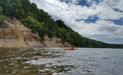 A person in a red kayak paddles on a calm river, with lush green cliffs and a cloudy sky in the background.