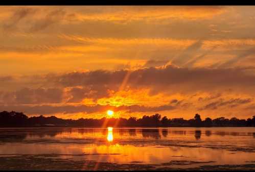 A vibrant sunset over a calm lake, with orange and yellow hues reflecting on the water and clouds in the sky.