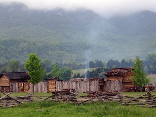 A rustic settlement with wooden structures and a smoky background, set against green mountains and a cloudy sky.