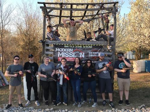 A group of people posing with toy guns in front of a wooden structure, smiling and enjoying an outdoor activity.