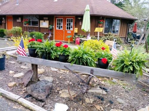 A rustic wooden planter with red flowers and ferns, flanked by small American flags, in front of a cozy brown building.