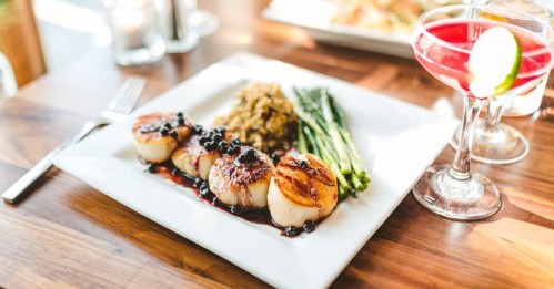 A plate of seared scallops with asparagus and risotto, accompanied by a cocktail on a wooden table.