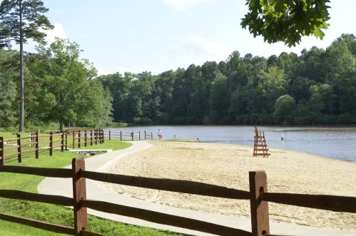 A sandy beach by a calm lake, surrounded by trees and a wooden fence, with a lifeguard stand in the background.