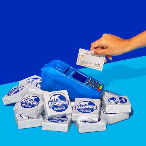 A hand holds a card above a blue payment terminal surrounded by Klondike ice cream bars on a colorful background.